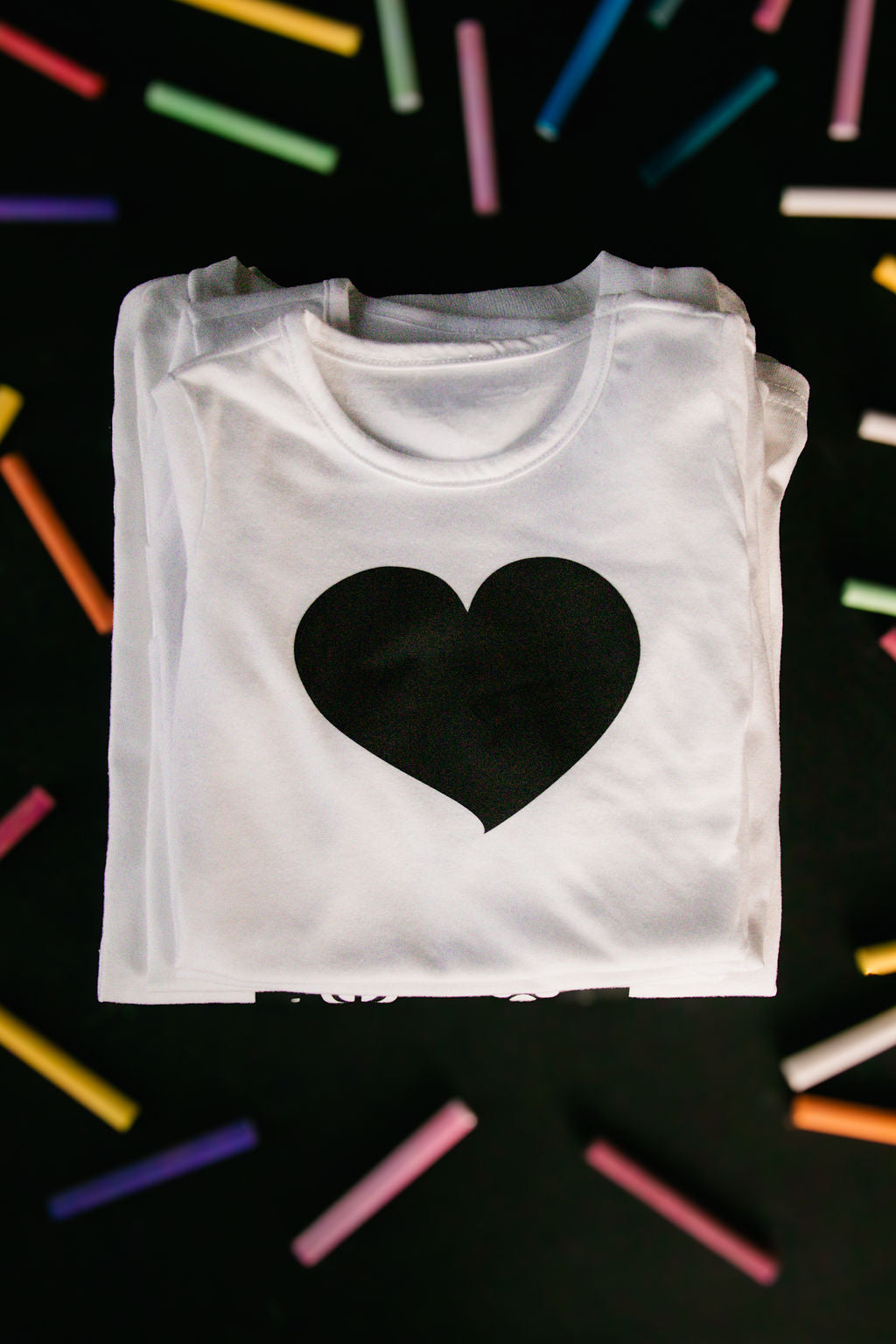 White t-shirt with a large black heart-shaped chalkboard vinyl design, neatly folded, surrounded by colorful chalk sticks on a black background.